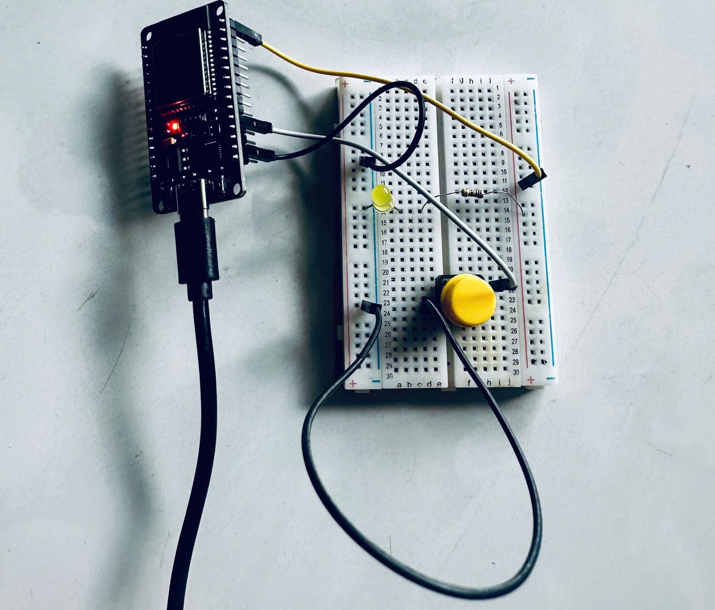 Ảnh chụp mạch thật: bo ESP32 DevKit cắm cáp USB, breadboard có LED vàng, điện trở và một nút nhấn vàng cỡ lớn.